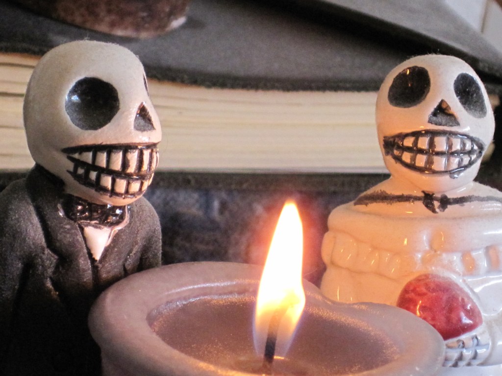small scuptures of skeletons in tuxedo and wedding dress in a Mexican folk art style, shown standing behind a lit candle and before a small stack of books.